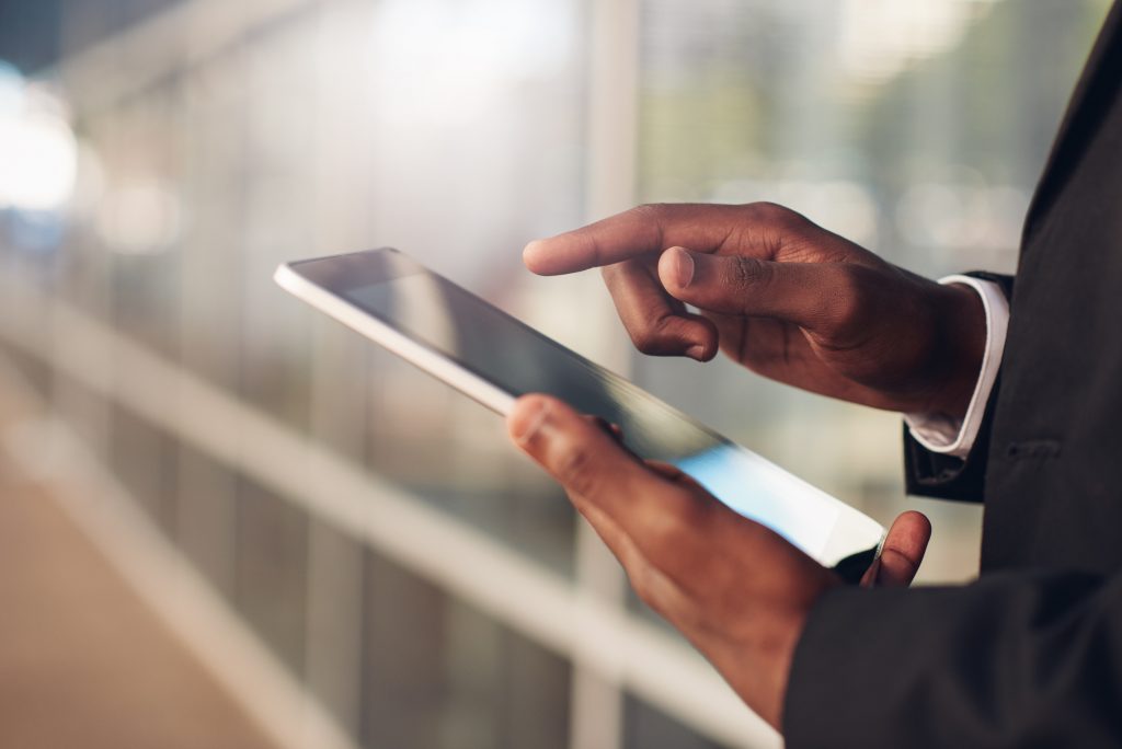 Cropped closeup of the hands of a businessman of African descent holding and using a digital tablet, wiht blurred office windows in the background