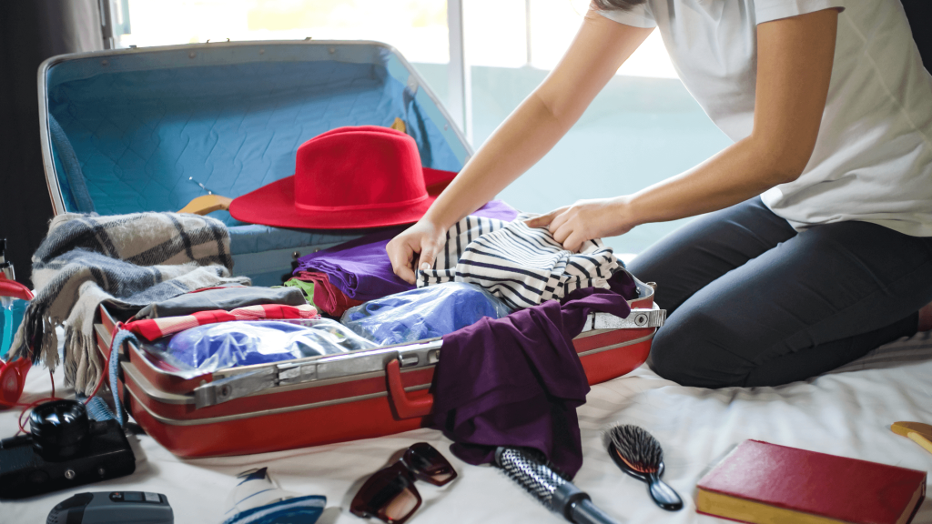 A woman sits on a bed, packing a suitcase filled with clothes. A travel CPAP, sunglasses, hairbrush, and other essentials sit on the bed with her.