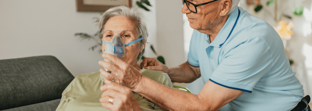 Senior man assisting senior woman during process of medical inhalation at home on the couch