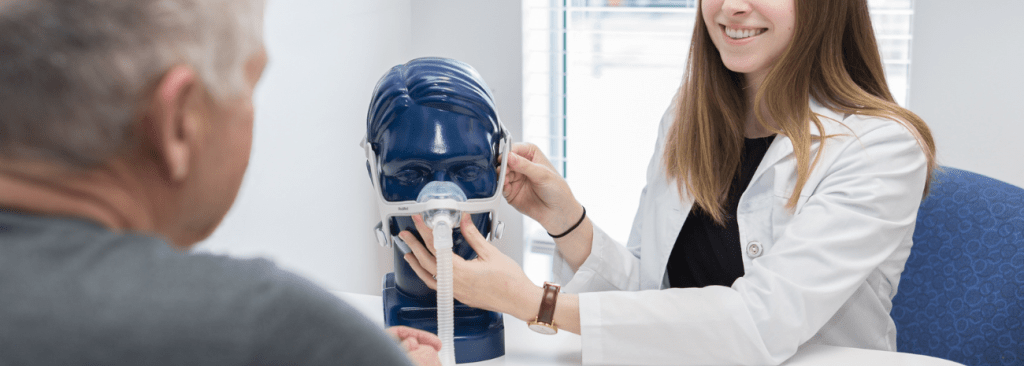 Healthcare professional in white coat instructing how to put CPAP mask on mannequin head to patient