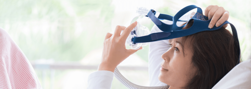 Woman lying in bed pulling a CPAP mask off of her head with a slight smile