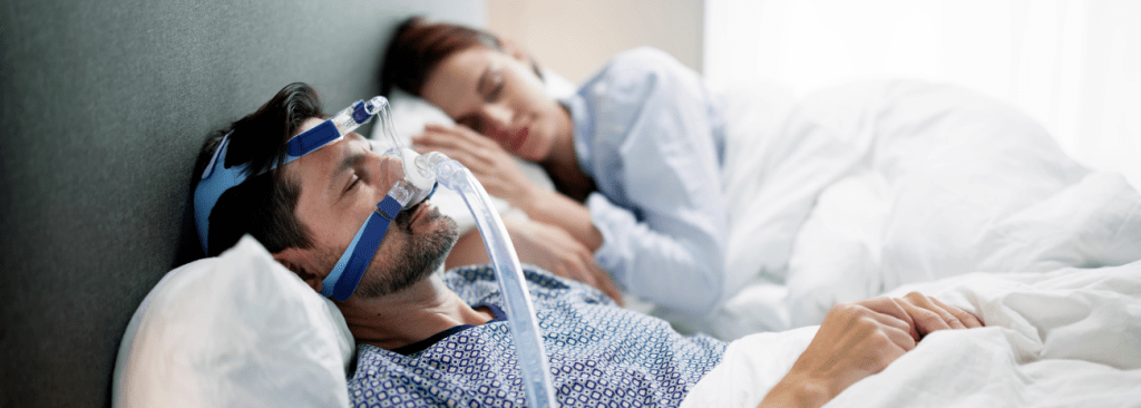Man with CPAP mask lying on bed next to woman sleeping