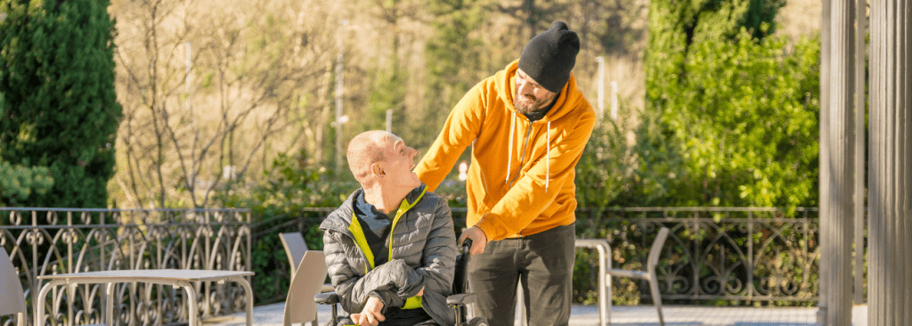Man pushing another man who is in a wheelchair while they are outside