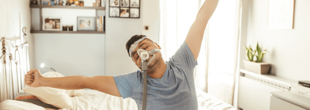 Man sitting on bed stretching his arms with a CPAP mask on his face