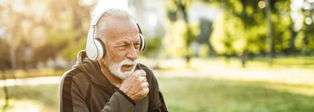 Sporty senior man walking outdoors with headphones on and coughing, out of breath