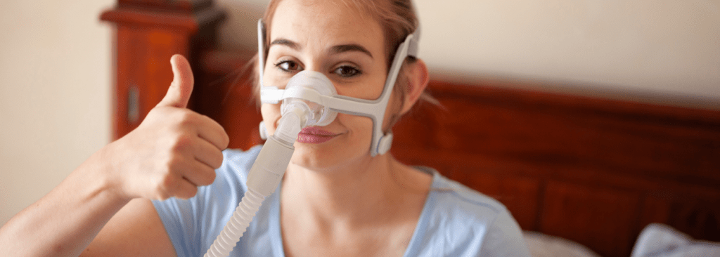 Woman sitting up in bed with a CPAP mask on giving a thumbs up and smiling