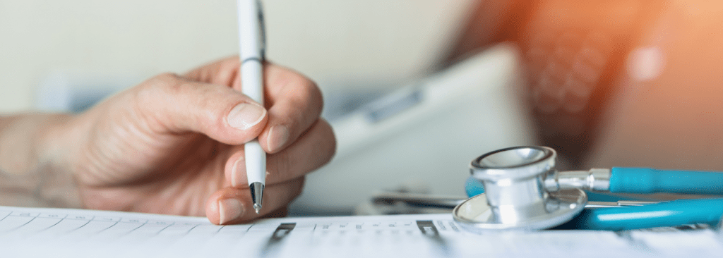 Healthcare professional writing with pen on discharge paperwork with telephone and stethoscope in background