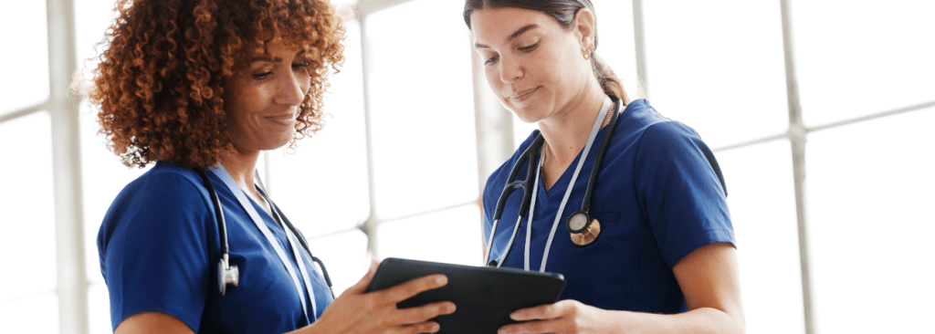 Two female healthcare professionals wearing scrubs and stethoscopes looking at tablet together and smiling
