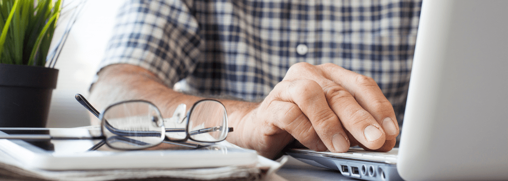 View of senior man's hands typing on laptop with his tablet and glasses next to him