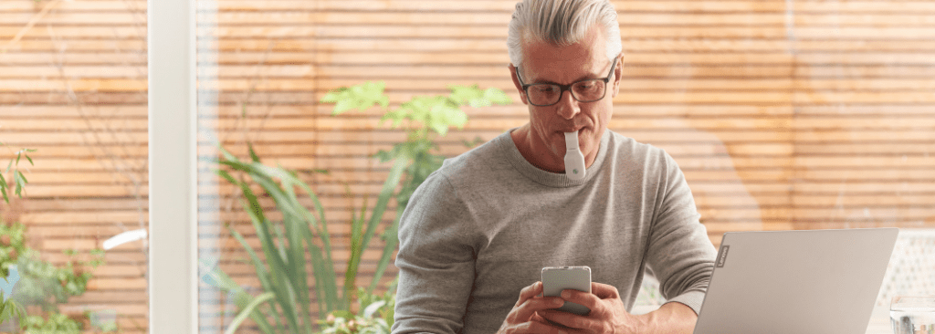 Man seated at table with eXciteOSA device in his mouth, while using app on his phone