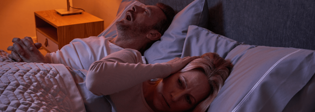 Man snoring in bed with wife covering her ears with her hands next to him