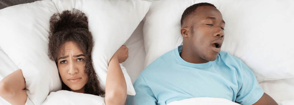 Woman pushing her pillow over her ears while man snores in bed next to her