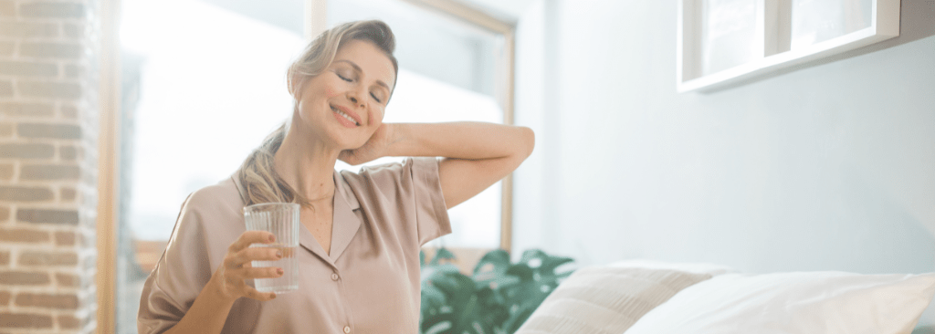 Woman sitting up in bed smiling with eyes closed, holding glass of water