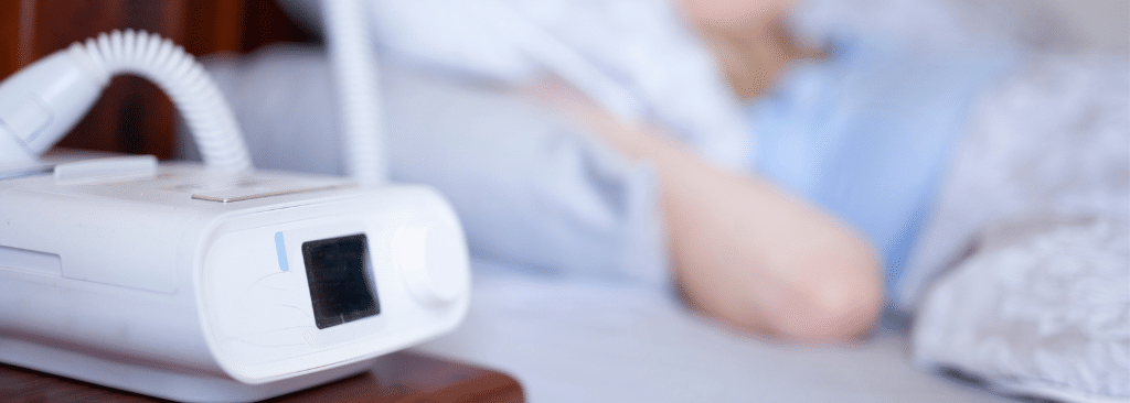 CPAP machine on bedside table with woman blurred out sleeping in the back with it on