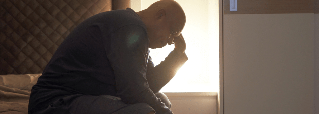 Older man sitting at the edge of the bed at night with his lamp on