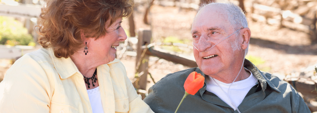 Senior man with oxygen cannula smiling at wife as they sit on bench outdoors