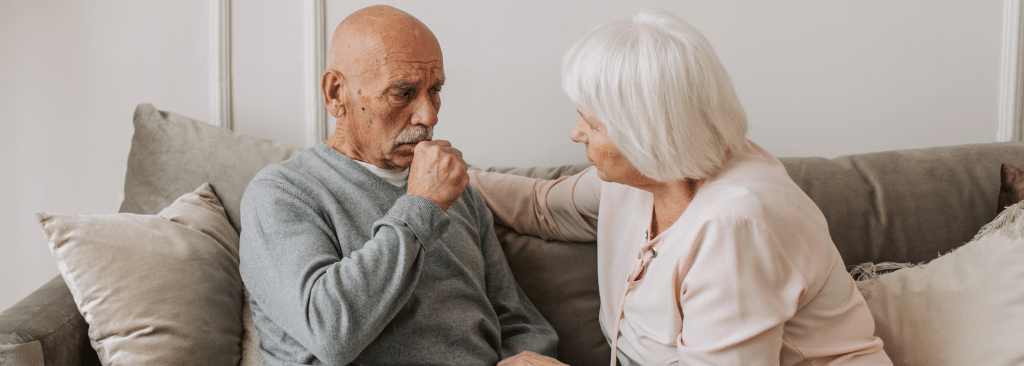 Older man sitting on couch with wife comforting him while he's coughing into his fist